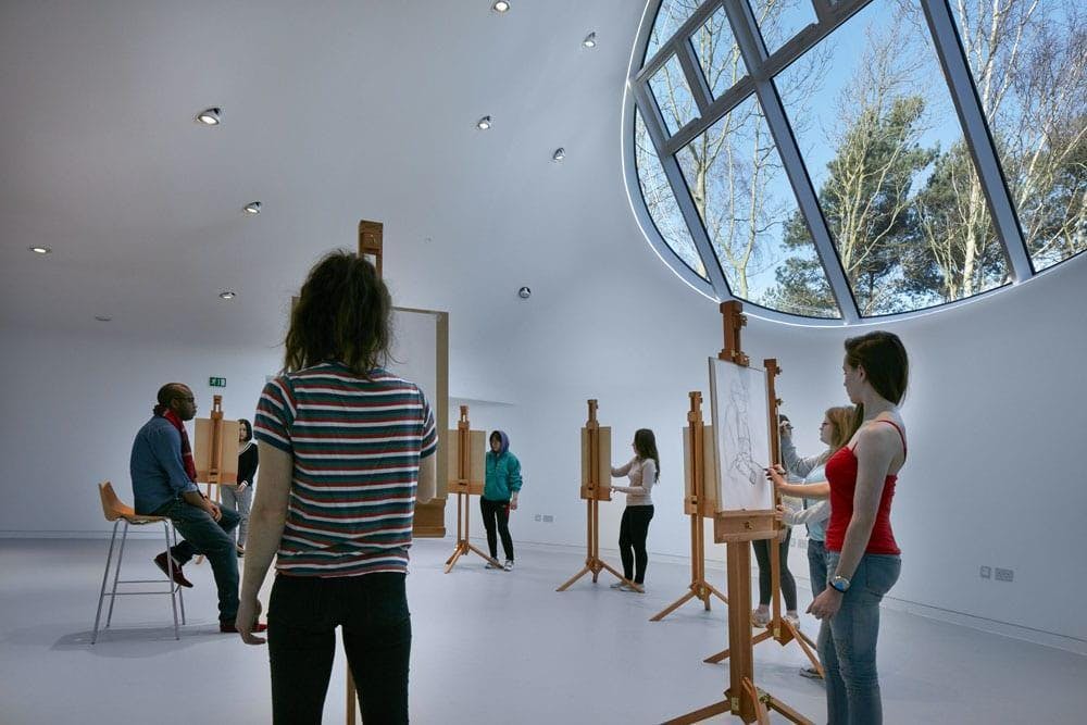 A model sits on a chair in the centre of a room while a group of artists draw them.