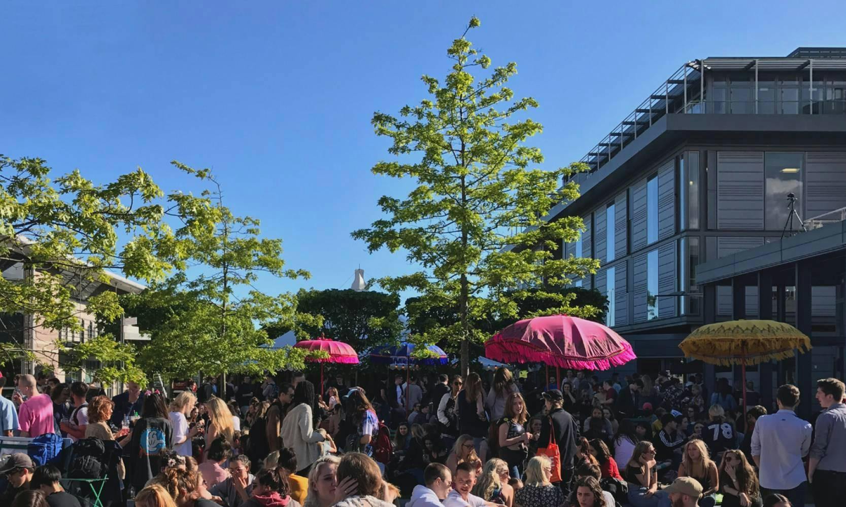 Closer image of a large crowd of people gather in a courtyard with trees and bright-coloured parasols in the background.