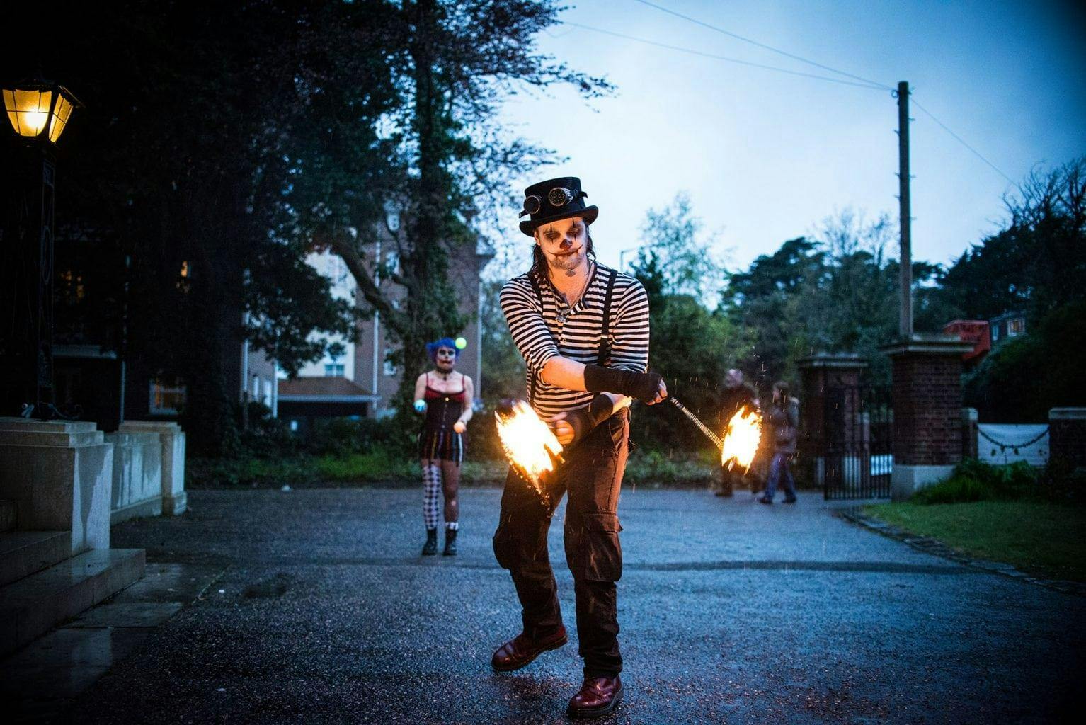 A fire juggler performing in costume with fire sticks on a pavement outside a building.