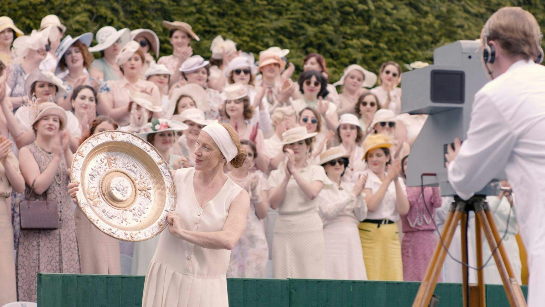 Performers in a period film. An actress in costume as a tennis player holds a gold shield trophy. A crowd of people in period costume are stood behind them, applauding.