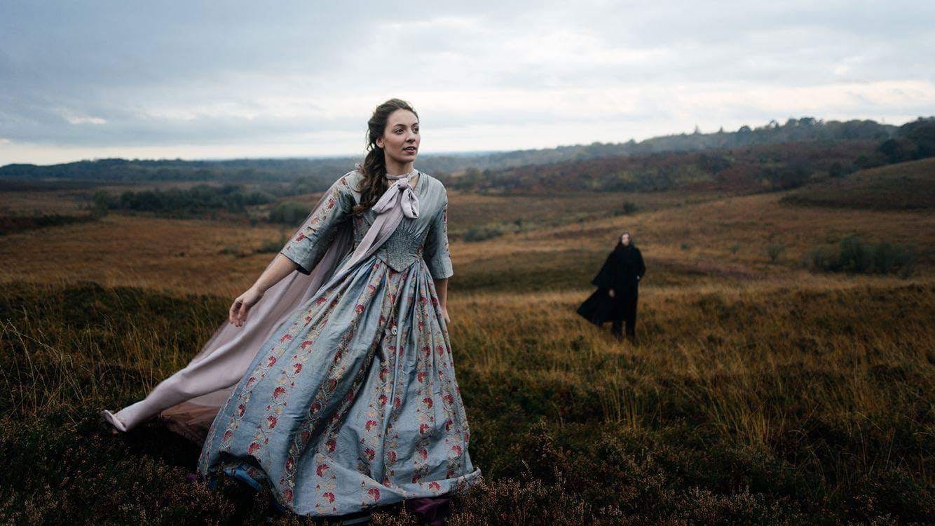A woman in period costume walking across a moor. A man, also in period costume, stands in the background and watches.