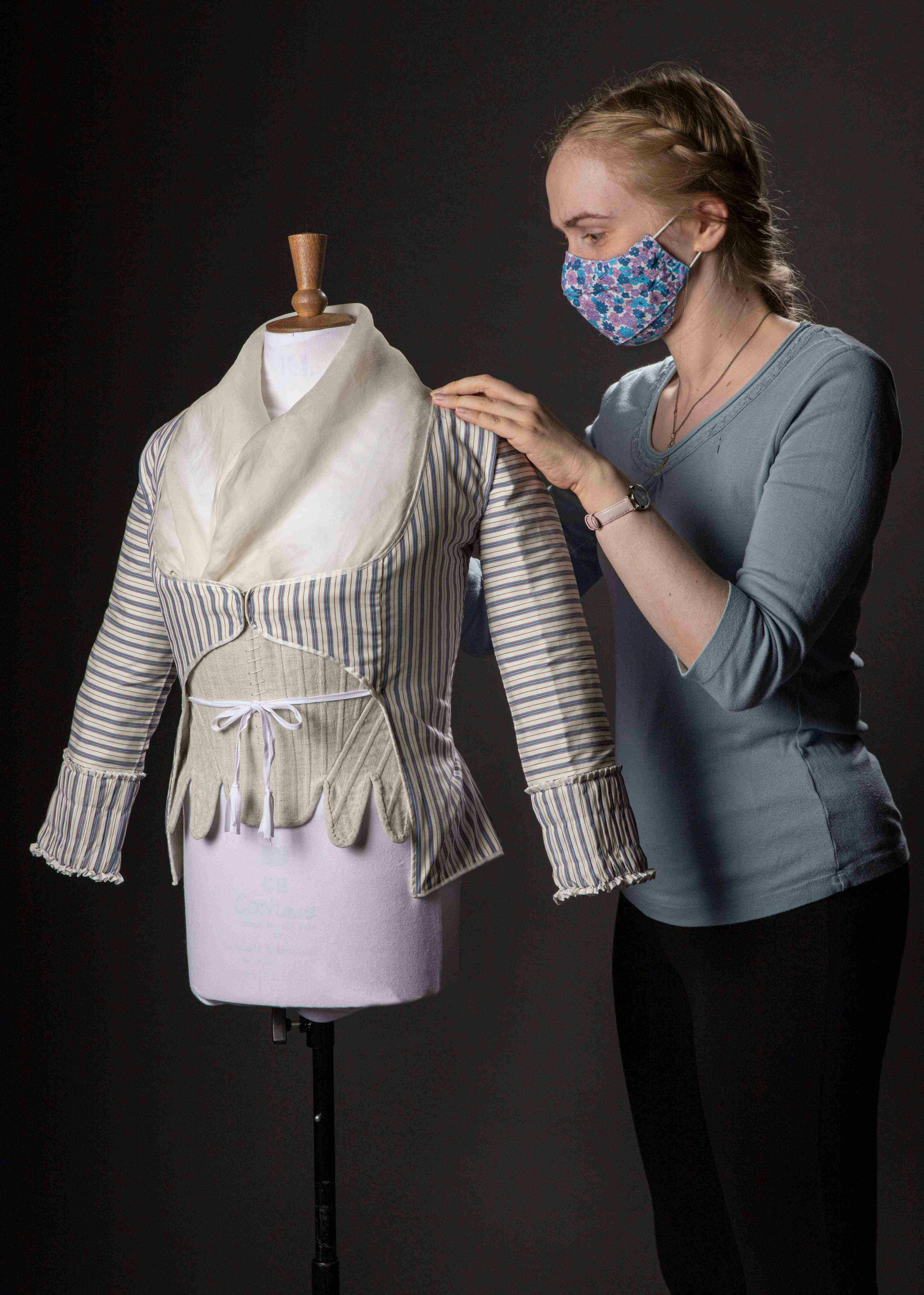 A person wearing a face mask works on a historical-style blouse on a mannequin.