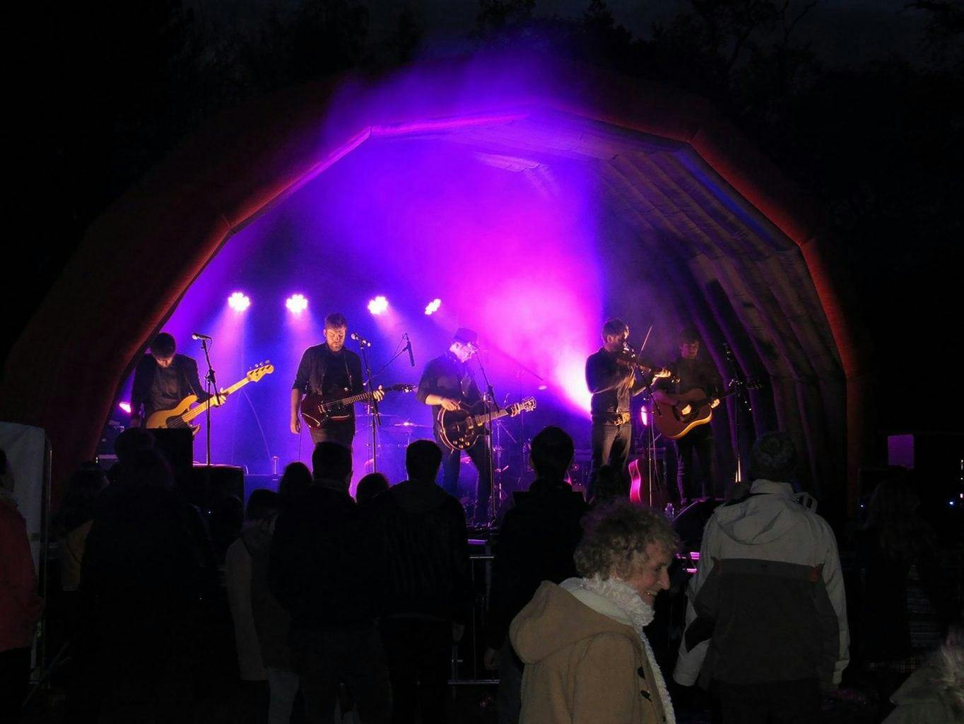A band on a stage lit with purple lights with a crowd in front.
