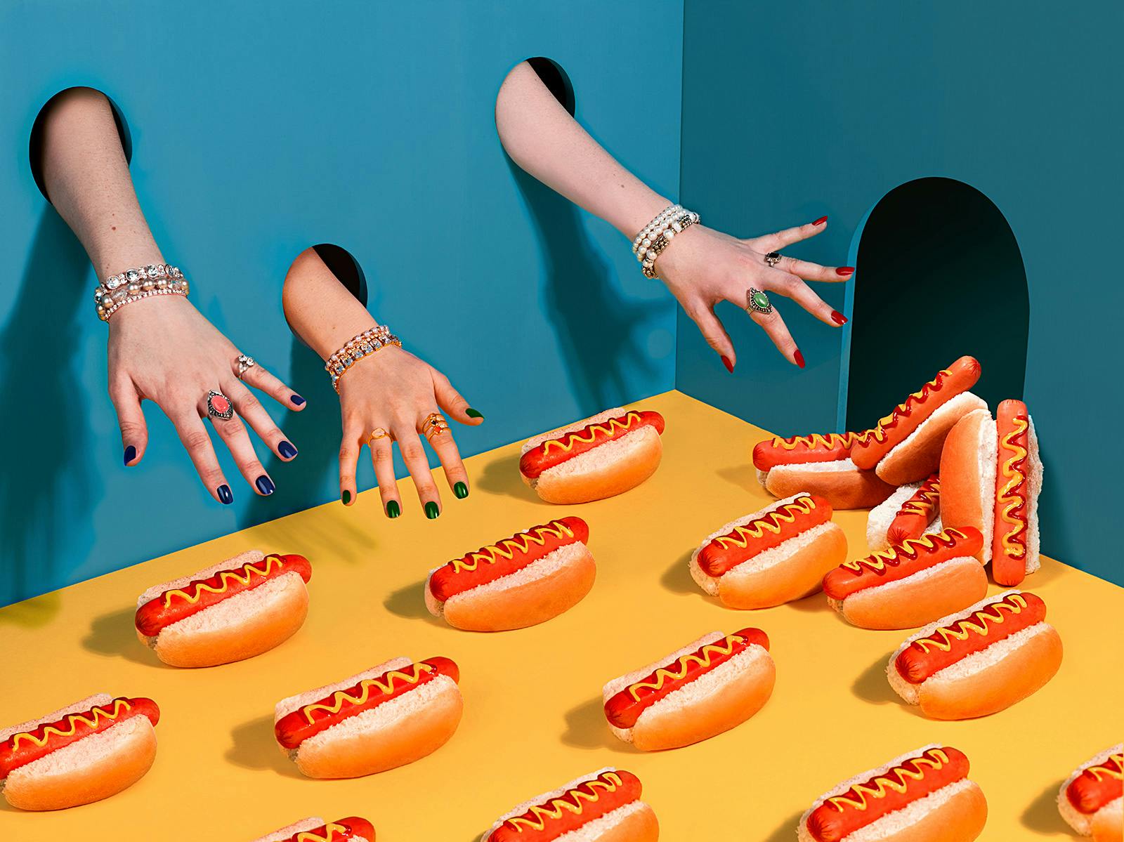 hands poking in through holes reaching for a pile up of hotdogs with mustard