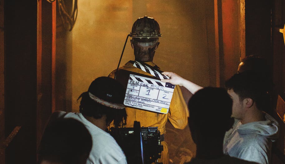 behind the scenes shot from the film Coal Miners. A man in mining gear is viewed from behind, while a camera man and another person holding a clapper board stand behind him.