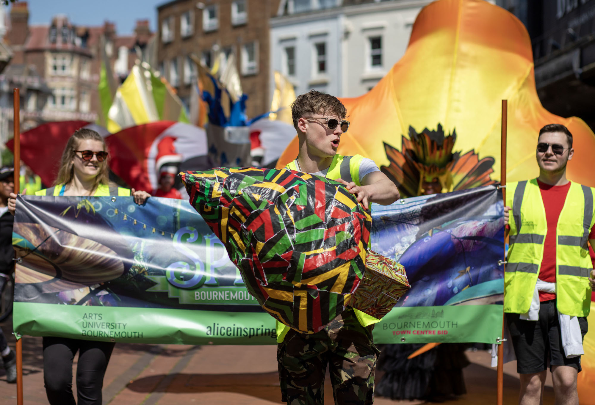 AUB student in hi-vis at the front of a parade holding a tree-shaped sculpture made of red, gold and green patterned paper.