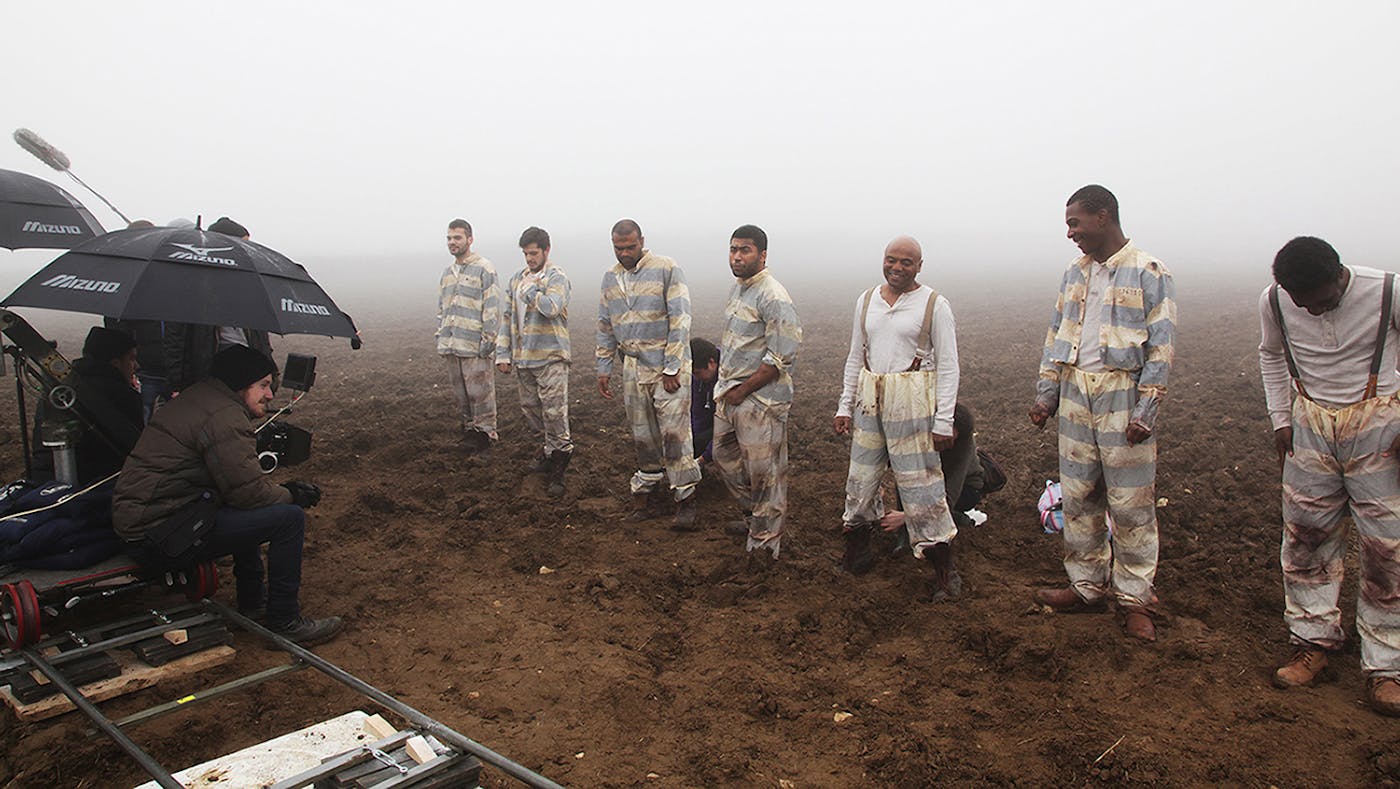 Actors in grey and white prisoner jumpsuit in a line, with a camera on a dolly track