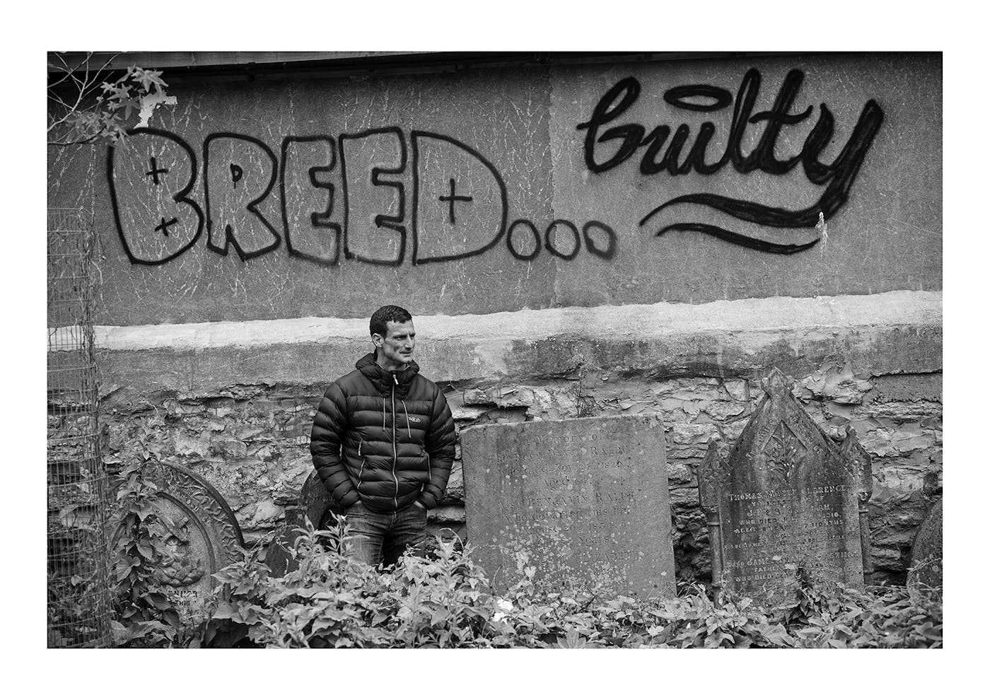 Black and white image of a man stood in front of a graffiti wall