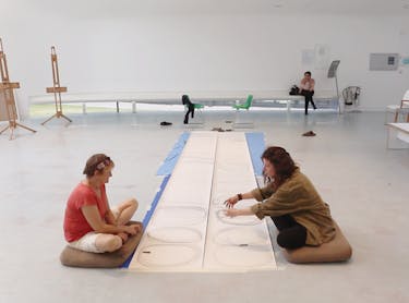 Two women sat on the floor with a long sheet of white paper in between them with circles drawn on.