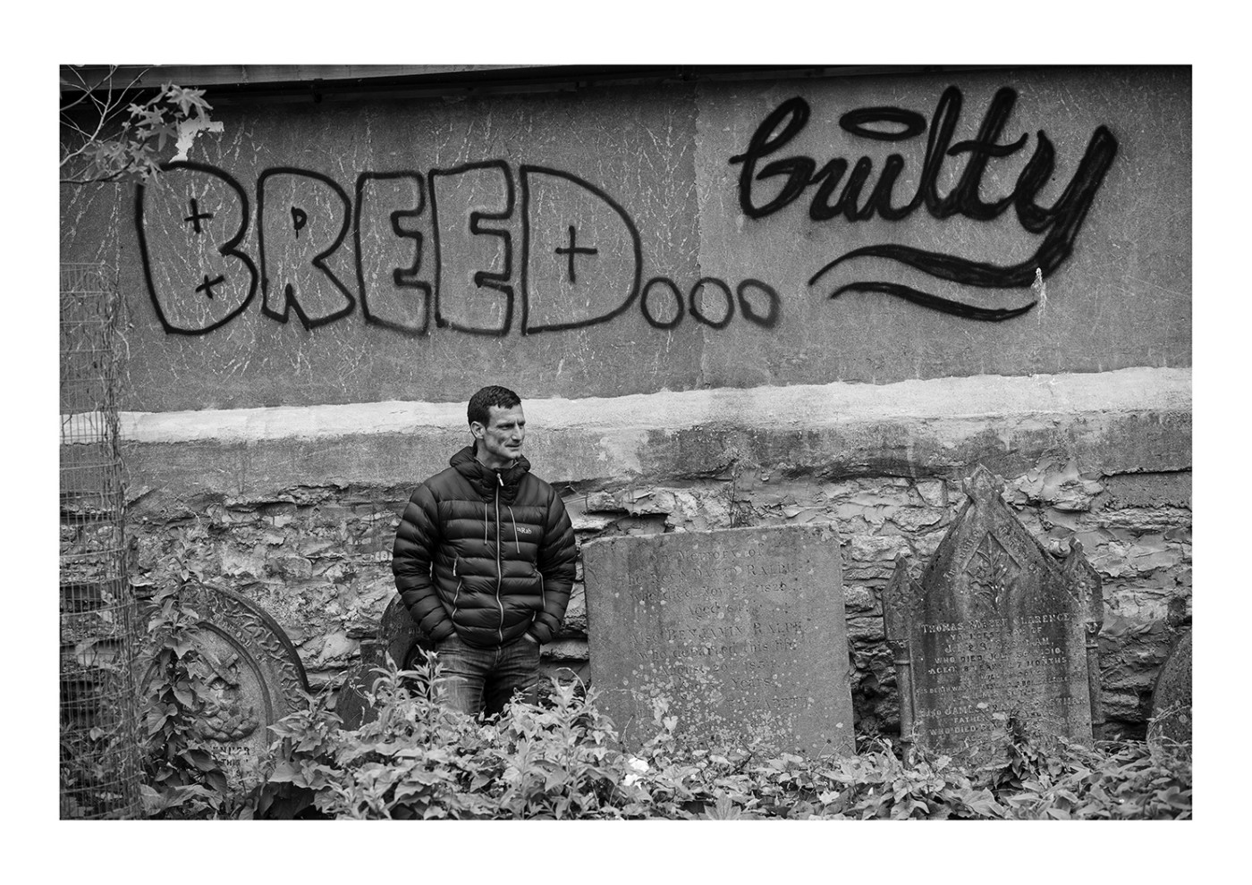 Black and white image of a man stood in front of a graffiti wall