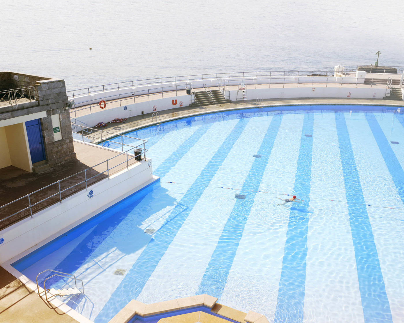 An open-aired swimming pool by the sea with a solitary elderly female swimmer