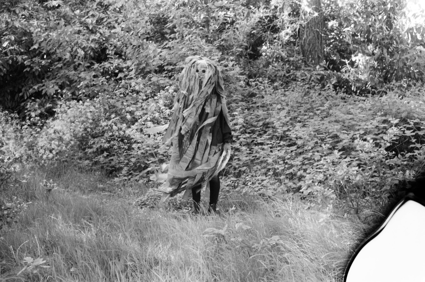 Black and white image of a women in a grassy field dressed head to toe in a costume made from long strips of fabric
