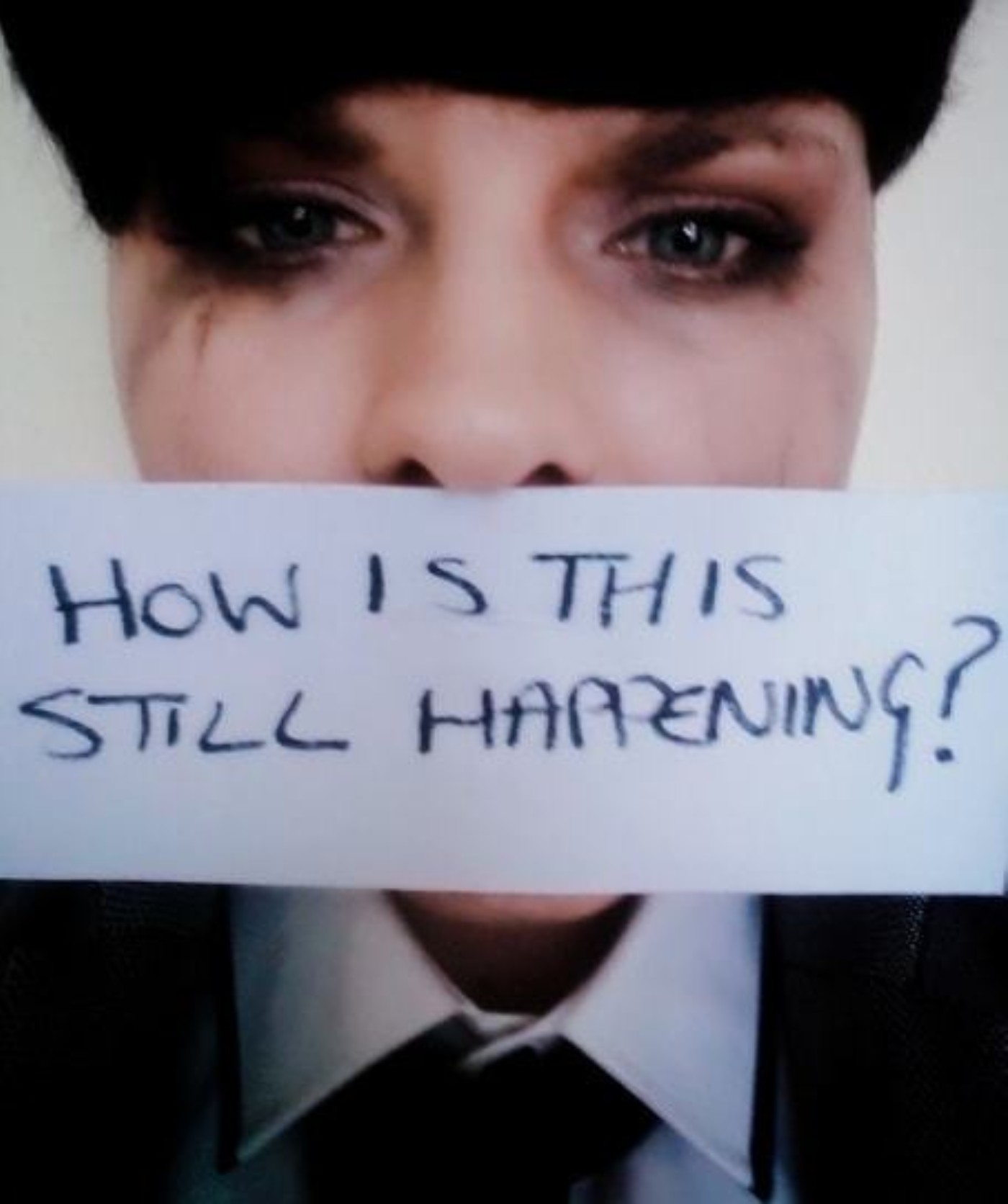 A person with smudged eye makeup holding a piece of paper reading 'HOW IS THIS STILL HAPPENING?' covering their mouth.