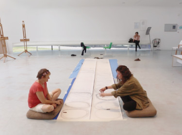 Two women sat on the floor with a long sheet of white paper in between them with circles drawn on.
